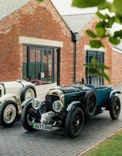 Two vintage Bentley Blower Jnr's, one dark green and one cream, parked on a brick driveway in front of a modern red brick building.
